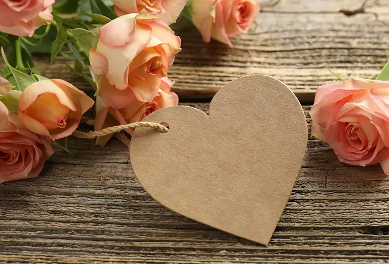 Herzförmiger Geschenkanhänger aus Kraftpapier mit Kordel, dekoriert mit rosafarbenen Rosen auf rustikalem Holzuntergrund.