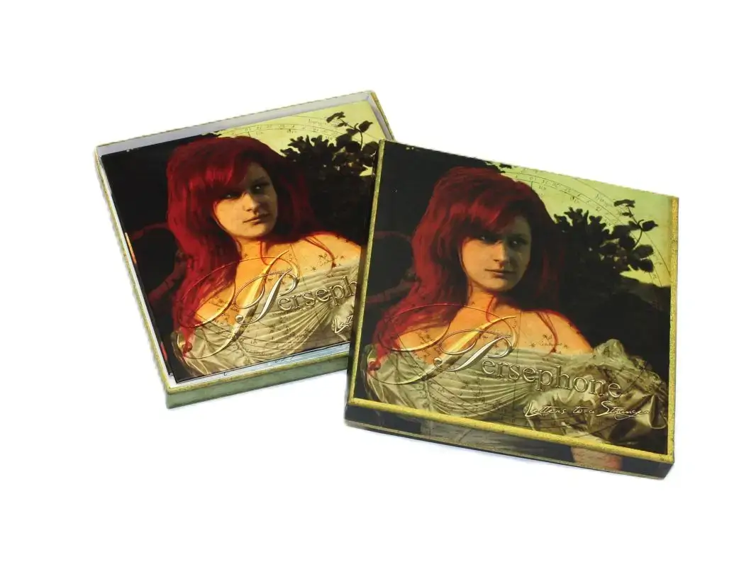 Opened square box in golden green with slanted lid and inserted booklet with cover motif of a woman with red hair in a light dress against a dark background.