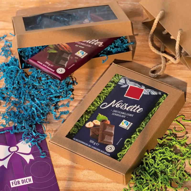 Brown gift boxes with window and chocolate bars on a wooden surface with colored filling material.
