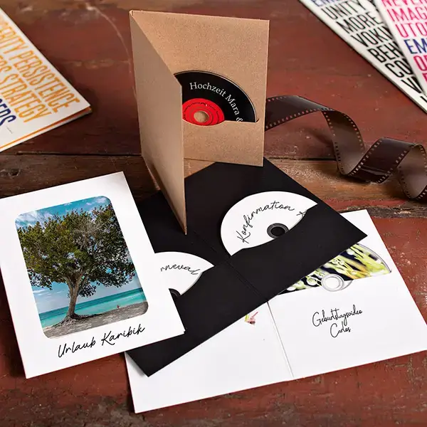 Geöffnete CD-Verpackungen aus Kraftpapier und schwarzem Karton mit eingelegten CDs und Fotos auf Holzuntergrund.