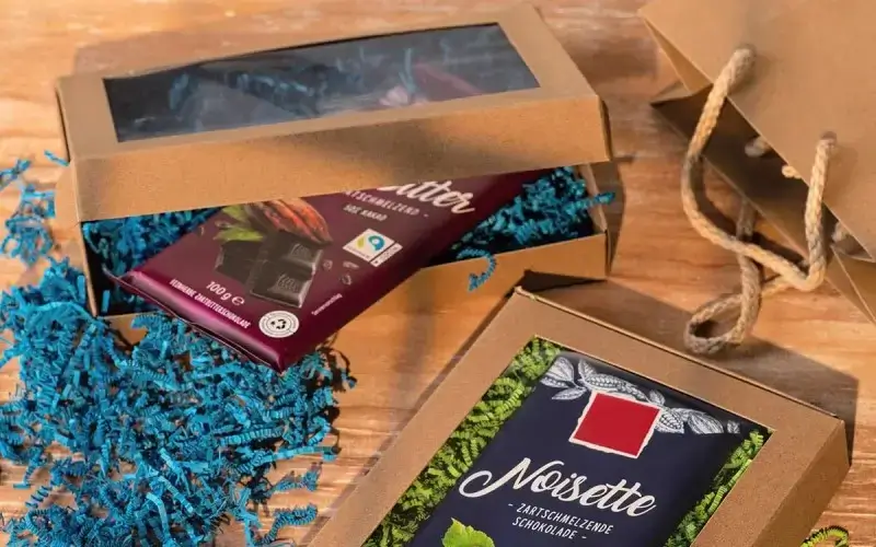 Kraft cardboard gift box with window and chocolate bar, decorated with blue paper filler on a wooden table.