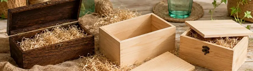 Trois caisses en bois teintées en clair et en foncé, posées sur une table rustique en bois, garnies de copeaux de bois, de toile de jute, de lierre et d'un verre à eau vert, dans une ambiance DIY lumineuse.